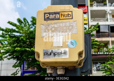 Scatola gialla in metallo montata su un palo. Superficie intemprata con adesivi sbiaditi. Fogliame verde visibile sullo sfondo. Ambiente urbano con edifici e cielo. Foto Stock