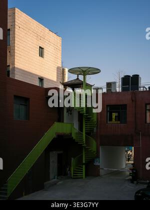 Wuhan, Cina - 22 marzo 2025: Scala a spirale in metallo verde attaccata all'esterno di un edificio industriale rosso e bianco. L'immagine mostra un urbano A. Foto Stock