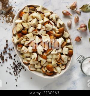 Funghi porcini freschi a fette in un piatto bianco su fondo in marmo. Gli ingredienti per la marinata sono sale, pepe nero, alloro, aglio, dil Foto Stock