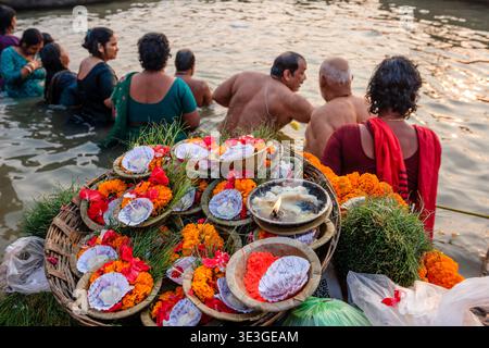 Un cestino di offerte di calendula e candele si trova in primo piano mentre la gente esegue il bagno rituale nel fiume Gange presso i ghat Varanasi. Foto Stock