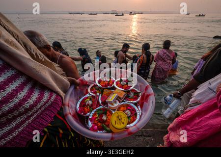 Una persona ai ghati di Varanasi tiene un vassoio di offerte rituali tra cui fiori e incenso mentre i devoti fanno il bagno nel fiume Gange. Foto Stock