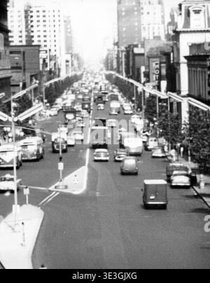 Questa fotografia aerea in bianco e nero di Angelo Rizzuto cattura il traffico stradale a due vie e l'architettura di una delle principali arterie di New York durante gli anni '1950 L'immagine mostra file di auto, autobus e veicoli dell'inizio della metà del secolo che si muovono lungo la strada, fiancheggiate da alti edifici cittadini. Foto Stock