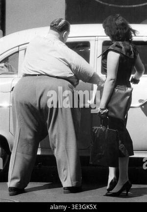 Dagli anni '1950, questa fotografia di strada in bianco e nero di Angelo Rizzuto cattura un momento della vita quotidiana a New York. Un uomo apre la porta di una macchina per una donna, mostrando la moda vintage e le classiche scene di strada dell'epoca. Foto Stock