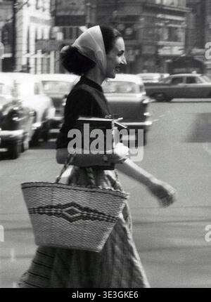 Una candida foto di strada scattata da Angelo Rizzuto, probabilmente degli anni '1950 o dei primi anni '1960, raffigura una donna elegante che cammina attraverso una strada della città, forse a New York. Porta con sé una borsa a cestino intrecciata e libri, che esemplificano la vita quotidiana e la moda dell'epoca. Foto Stock