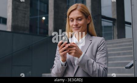 Sorridente felice donna d'affari caucasica che sfoglia e-mail al telefono in città utilizzando un'imprenditrice di servizi mobili lavoratrice donna in centro vicino all'ufficio Foto Stock