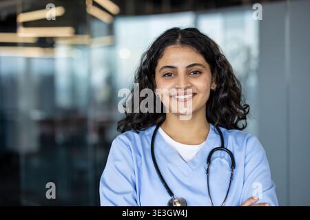 Sorridente giovane donna indiana adulta che indossa scrub azzurri e uno stetoscopio, in piedi con sicurezza con le braccia incrociate in un moderno ambiente di studio medico Foto Stock