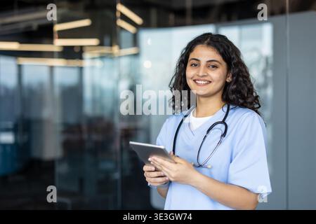 Giovane professionista medico che indossa scrub blu e uno stetoscopio, in piedi in un ufficio e utilizzando un tablet digitale per la tecnologia sanitaria e la cura dei pazienti Foto Stock