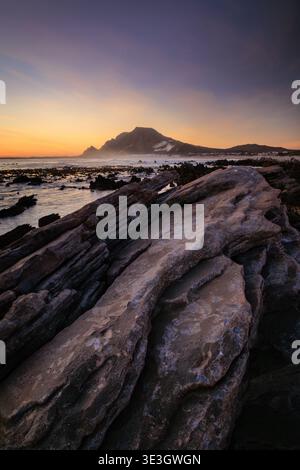 Vista delle aspre formazioni rocciose si estendono verso il mare sotto un vivace cielo crepuscolo, con una montagna lontana che si staglia contro il caldo orizzonte, Bet Foto Stock