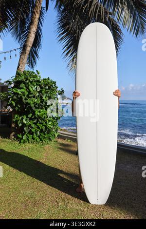 Uomo in possesso di una tavola da surf bianca su una spiaggia tropicale con vista sull'oceano Foto Stock