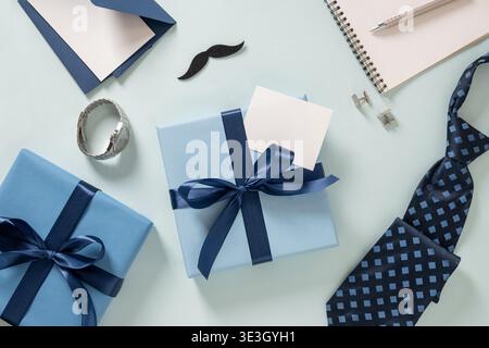 Composizione creativa per la festa del papà con regali avvolti, cravatta a motivi geometrici, gemelli e bigliettino vuoto disposti su sfondo blu pastello. Stile dall'alto. Copia spazio. Foto Stock