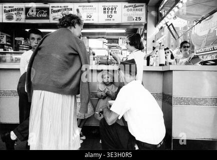 Questa accattivante fotografia di strada, probabilmente catturata da Angelo Rizzuto negli anni '1950, mostra le persone riunite intorno a un vivace bancone di ristoranti di New York City. Adulti e bambini, vestiti in stile d'epoca, interagiscono mentre i tabelloni dei menu pubblicizzano piatti americani classici come hamburger e frkfurter, offrendo uno sguardo sulla vita quotidiana in un vivace ambiente urbano. Foto Stock