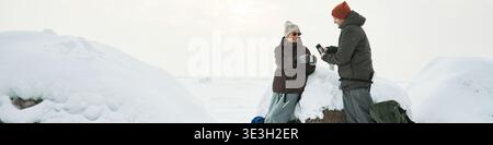 Intestazione di donna caucasica e uomo di mezza età in piedi all'aperto su un paesaggio innevato sorridendo e tenendo in mano tazze, impegnandosi in conversazione durante l'avventura invernale, rocce innevate visibili Foto Stock