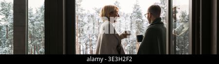 Intestazione di una donna caucasica di mezza età e di un uomo caucasico di mezza età in piedi accanto alla finestra tenendo tazze, impegnandosi in una conversazione con la foresta innevata visibile all'esterno, entrambi avvolti in coperte Foto Stock