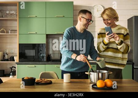 Uomo di mezza età caucasico che mescola la pentola su una stufa sorridendo, accanto a una donna di mezza età caucasica che tiene in mano lo smartphone e ride insieme in un moderno interno della cucina Foto Stock