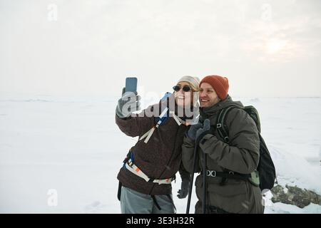 Uomo e donna caucasica di mezza età in piedi all'aperto in un paesaggio innevato, sorridendo e scattando selfie con lo smartphone indossando zaini e cappelli invernali Foto Stock