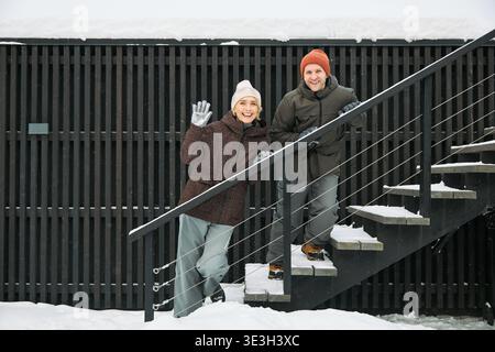 Donna caucasica di mezza età che sventola e sorride mentre sta in piedi su una scala innevata all'aperto accanto a un uomo caucasico di mezza età sorridente, entrambi indossano cappelli e guanti invernali in ambiente invernale Foto Stock