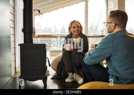 Donna caucasica di mezza età sorride e tiene in mano la tazza mentre siede vicino a una stufa a legna, parla con un uomo caucasico di mezza età, entrambe rilassanti all'interno in un'accogliente cabina Foto Stock