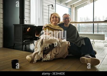Donna caucasica di mezza età e uomo caucasico adulto seduto su un pavimento di legno vicino al caminetto, sorridente e avvolgente avvolto da coperte, guardando la macchina fotografica nell'accogliente interno della cabina Foto Stock