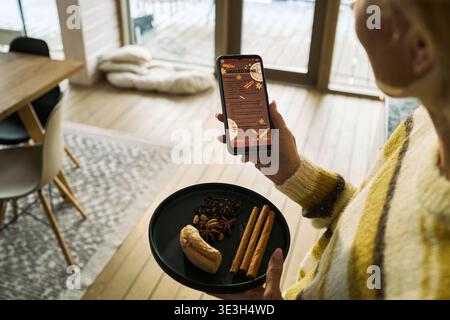 Donna caucasica di mezza età che tiene in mano uno smartphone che mostra la ricetta del VIN brulé mentre trasporta un piatto nero con bastoncini di cannella, anice stellato, chiodi di garofano e zenzero nella moderna cucina casalinga Foto Stock