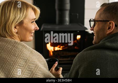 Donna caucasica di mezza età sorridente e tiene in mano una tazza mentre siede accanto a un uomo caucasico di mezza età, entrambe rilassandosi insieme davanti al caminetto e godendo di una calda atmosfera Foto Stock