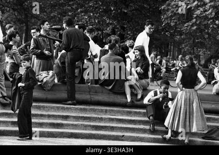 Una candida foto di strada in bianco e nero di Angelo Rizzuto, scattata nell'autunno del 1953, mostra un vivace raduno di persone a Washington Square, New York City. Gli individui si vedono seduti su gradini e sporgenze, chiacchierare e suonare strumenti musicali, riflettendo la moda casual quotidiana e la scena sociale dell'epoca. Foto Stock