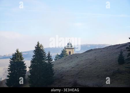 Piccola cappella con tetto a cupola in piedi su una collina ghiacciata circondata da alberi in un paesaggio rurale Foto Stock