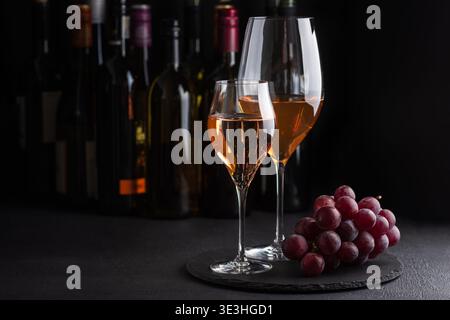 Due bicchieri di vino bianco e di rosa frizzante con grappoli di uva su una superficie scura di ardesia su bottiglie sullo sfondo Foto Stock