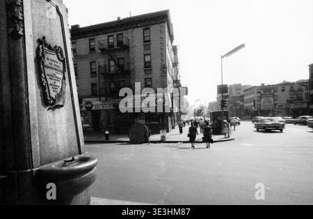 Questa fotografia di strada in bianco e nero degli anni '1950 di Angelo Rizzuto cattura la vivace intersezione tra Delancey Street e Bowery a New York City. I pedoni percorrono la tipica moda degli anni '1950 sullo sfondo di vetrine, auto d'epoca e la monumentale fontana Timothy P. Sullivan. Foto Stock