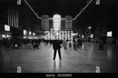 Questa accattivante fotografia in bianco e nero, probabilmente di Angelo Rizzuto degli anni '1940 o '1950, cattura l'animato interno del Grand Central Terminal di New York City. Una figura solitaria si erge con le spalle allo spettatore, osservando il turbinio dell'attività quotidiana e la grandiosa architettura dell'atrio, che include grandi finestre ad arco e un'importante insegna "HI-FI HOUSE". Foto Stock