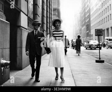 Questa accattivante fotografia di strada in bianco e nero di Angelo Rizzuto, probabilmente degli anni '1950, cattura una coppia elegante che si tiene per mano mentre cammina lungo un vivace marciapiede di New York. Il cappello a teglia larga da donna, la top a righe e la gonna a pieghe incarnano le tendenze di moda dell'epoca, contrastando con l'abito affilato e gli occhiali da sole scuri del suo compagno maschile, creando una classica scena vintage di vita urbana. Foto Stock