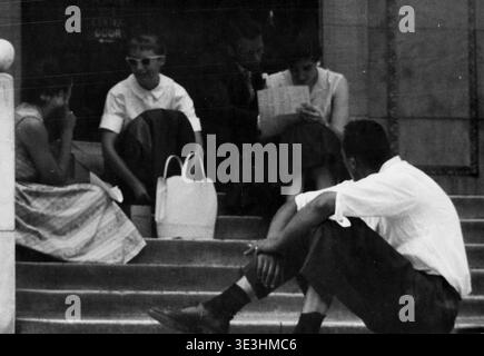 Questa fotografia di strada vintage, probabilmente scattata da Angelo Rizzuto negli anni '1950, mostra un gruppo di giovani seduti casualmente sui gradini di un edificio a New York. Sono vestiti con la tipica moda degli anni '1950, impegnati in attività quotidiane come la lettura e la conversazione, riflettendo lo stile schietto della fotografia di strada di quell'epoca. Foto Stock