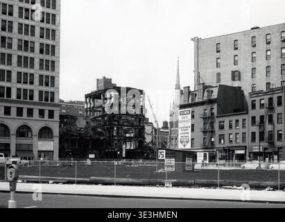 Questa fotografia in bianco e nero degli anni '1950 cattura una sezione di New York, mostrando le conseguenze dell'incendio dell'edificio Wanamaker. A destra, edifici intatti con un cartello "David Schwartz" per la pelletteria e i bagagli, e un campanile della chiesa, contrastano con i resti scheletrici di una struttura bruciata, evidenziando la vita quotidiana in mezzo al cambiamento urbano. Foto Stock