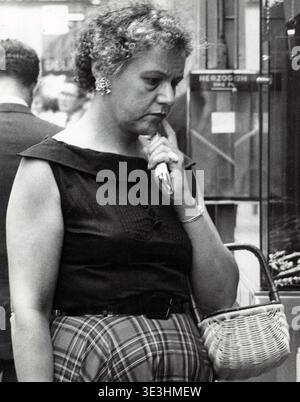 Questa accattivante fotografia di strada di Angelo Rizzuto, probabilmente degli anni '1950, cattura una donna di New York con un'espressione premurosa, tenendo in mano un accendisigari. È vestita in modo appropriato al periodo, con un top senza maniche scuro con un ampio colletto e una gonna tartan o a quadri, che riflette lo stile quotidiano della vita urbana della metà del secolo. Foto Stock