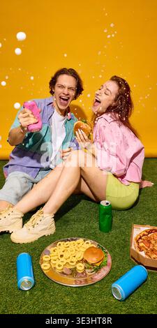 Felice giovane coppia seduta sull'erba a ridere con le lattine di fast food e bevande. Foto Stock