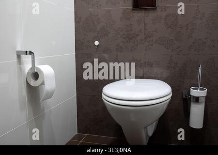 Moderna ciotola in ceramica bianca montata su una parete piastrellata scura in un bagno pulito con un rotolo di carta igienica Foto Stock