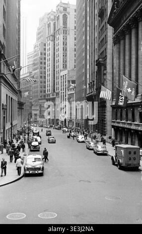 Questa fotografia vintage di strada in bianco e nero di Angelo Rizzuto, probabilmente degli anni '1950, cattura una vivace scena che si affaccia su Broad Street da Wall Street a New York City. I pedoni in stile d'epoca riempiono i marciapiedi accanto a numerose auto d'epoca, offrendo uno sguardo sulla vita quotidiana nel quartiere finanziario. Foto Stock