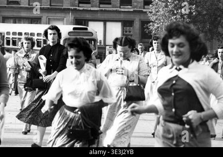 Questa accattivante fotografia di strada, probabilmente catturata da Angelo Rizzuto a metà degli anni '1950, raffigura una folla di donne in una strada di New York, probabilmente vicino al traghetto Staten Island. L'immagine mostra in modo splendido la moda di tutti i giorni dell'epoca, con vari stili di gonne, camicette e accessori portatili tipici della vita urbana degli anni '1950. Foto Stock