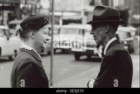 Questa fotografia in bianco e nero degli anni '1950 di Angelo Rizzuto cattura un momento sincero tra un uomo e una donna che conversano in una strada di New York. L'immagine mostra la moda tipica dell'epoca, con l'uomo che indossa una fedora e un abito, e la donna con un modesto cappello e un abito con colletto. Foto Stock
