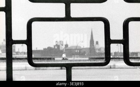 Questa fotografia in bianco e nero degli anni '1950 di Angelo Rizzuto cattura una vista sfocata dello skyline di New York, incorniciata da ornati lavori in ferro. Una figura lontana si siede su una panchina, aggiungendo un senso di vita quotidiana e un'introspezione urbana alla scena. Foto Stock