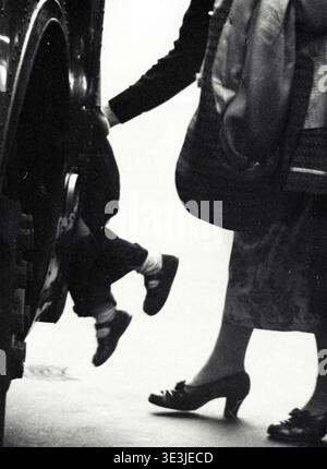 Questa accattivante fotografia di strada di Angelo Rizzuto, probabilmente degli anni '1940, cattura un momento comune della vita quotidiana a New York con una madre che guida un bambino su un autobus o un tram. La composizione si concentra intimamente sui loro piedi e sugli stili vintage delle loro scarpe, offrendo uno sguardo al passato. Foto Stock