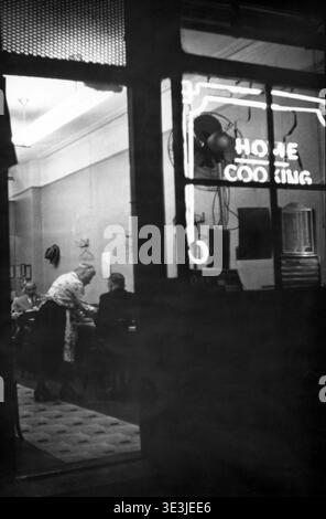 Questa accattivante fotografia di strada di Angelo Rizzuto, probabilmente degli anni '1940, offre uno sguardo su un ristorante di New York City. Girato attraverso una finestra di notte, mostra un ristorante all'interno con un cartello al neon che pubblicizza "CUCINA CASALINGA" e una cameriera che si occupa dei clienti, catturando un momento della vita quotidiana nella New York vintage. Foto Stock