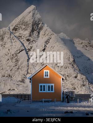 La vista di una calda casa gialla si erge in netto contrasto con lo sfondo innevato delle montagne, una serena scena invernale, Hamnøy, Nordland, Norvegia. Foto Stock