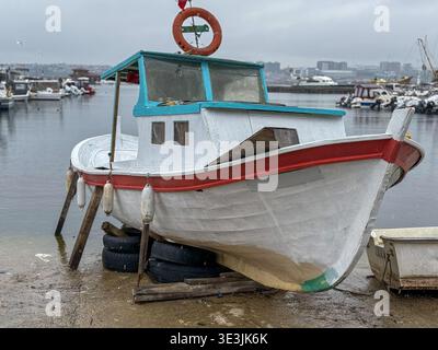 Vista di una barca da pesca bianca e rossa intemprata che poggia sulle gomme, vicino a un porto sotto un cielo nuvoloso, Sarıyer, İstanbul, Türkiye. Foto Stock