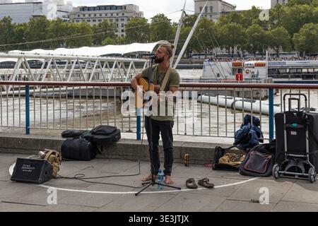 Musicista di strada che suona la chitarra acustica su una passeggiata sul lungofiume della città. Londra, Regno Unito, 23 luglio 2023 Foto Stock