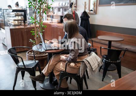 Donna che lavora su un computer portatile e beve un caffè al grande tavolo da caffè con finestra e luce solare. Pausa caffè, laptop, luce solare, tavolino, donna che lavora. Foto Stock