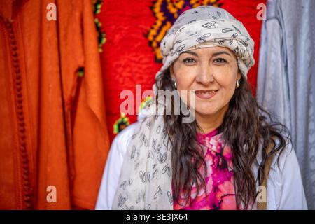 Una donna sorridente che indossa un velo a motivi geometrici e un abbigliamento luminoso si staglia davanti a tessuti vivaci in un mercato locale, trasmettendo calore, tradizione e.. Foto Stock