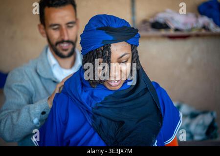 Donna sorridente che indossa un turban e una sciarpa blu in stile tuareg, mentre un uomo aiuta a regolare i suoi vestiti in un caldo villaggio marocchino, catturando Foto Stock
