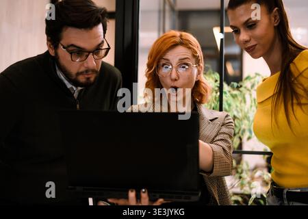Tre colleghi si riuniscono intorno a un notebook in un ufficio moderno, esprimendo sorpresa ed entusiasmo per una presentazione o per i dati sullo schermo durante un Foto Stock