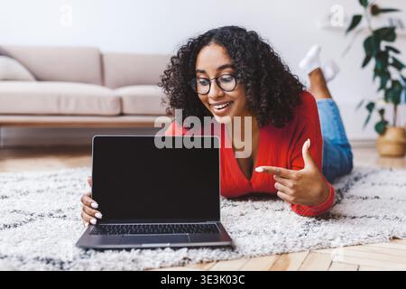 Una giovane donna si sdraia su un tappeto e punta sul laptop mentre sorride in una stanza luminosa Foto Stock