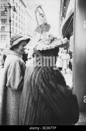 Questa accattivante fotografia di strada di Angelo Rizzuto, probabilmente degli anni '1950, cattura due donne su un marciapiede di New York. La donna in primo piano sfoggia un cappello straordinariamente elaborato ornato da un cestino e un coniglietto, suggerendo una celebrazione pasquale, mentre la donna accanto al suo sorride calorosamente. Foto Stock